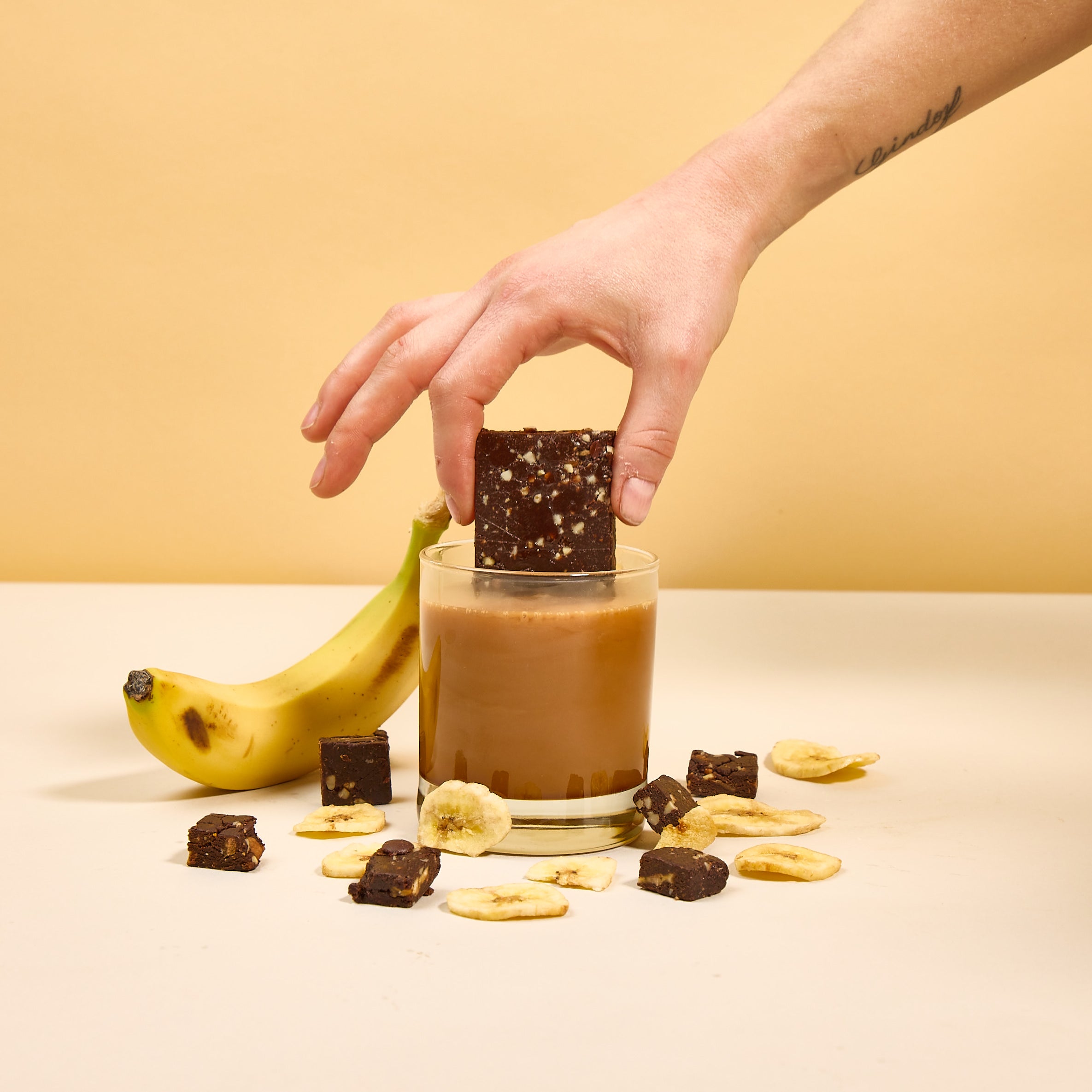 Hand holding Lucid's Banana Bread Snack Bar, dipping into a glass of caramel, with a banana leaning against the glass and surrounded by chocolate pieces and banana chips.