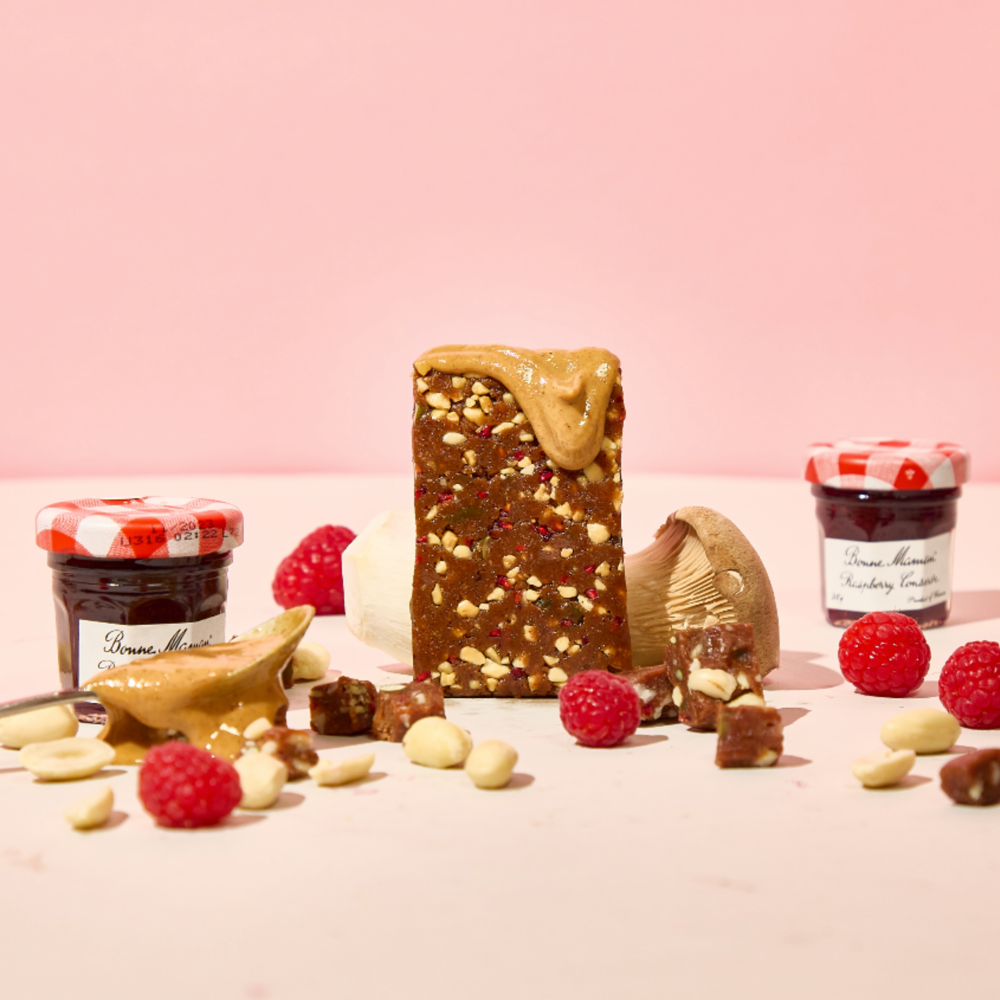 A Lucid Peanut Butter and Jelly bar with peanut butter and raspberries, alongside nuts, chocolate, mushrooms and two Bonne Maman jam jars, against a pink background.